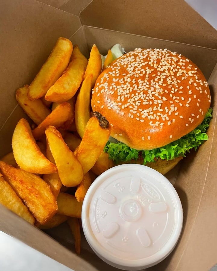 Takeaway box with thick cut golden crispy chops and burger, with green gem lettuce and sesame seeded bu. In corner is white polystyrene cup of sauce with white lid on top. Burton Farm, Local Farm shop, Chippenham.