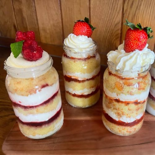 Three 'cakes in jars'; layers of sponge cake , jam and cream filling in a mason-style jar with whipped cream and strawberries and raspberries on top. Burton Farm, Local Farm shop, Chippenham.